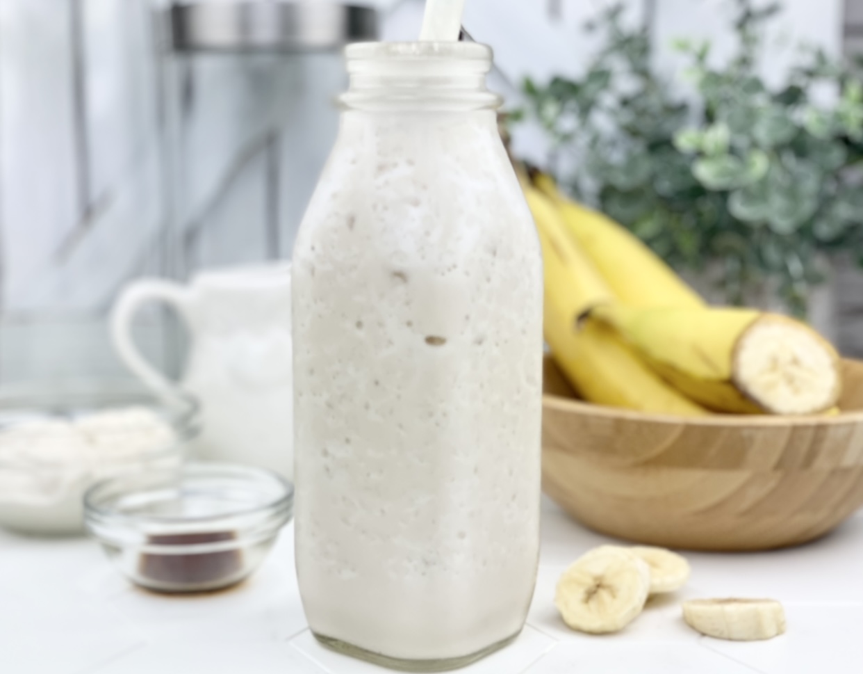 Photo of the creamy banana protein smoothie shwn in a glass with the ingredients surrounding it. 